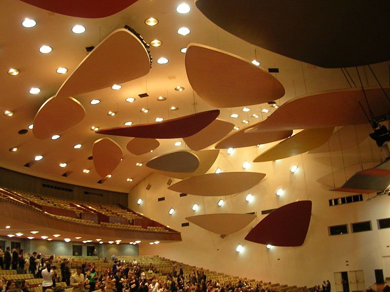 Curved coloured rafts suspended below the ceiling of a large auditorium, above rows of seating and an audience.