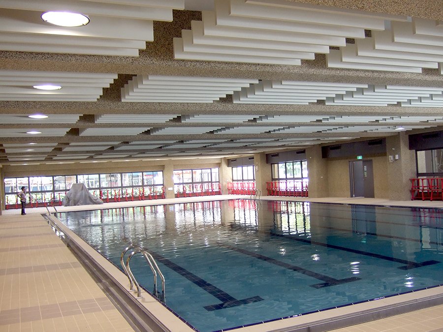 Rows of white baffles across the ceiling of an indoor swimming pool hall, above a tiled pool surround.