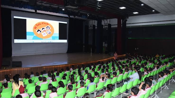 School hall full of children in green seats watching a projected image, with loudspeakers flanking the stage