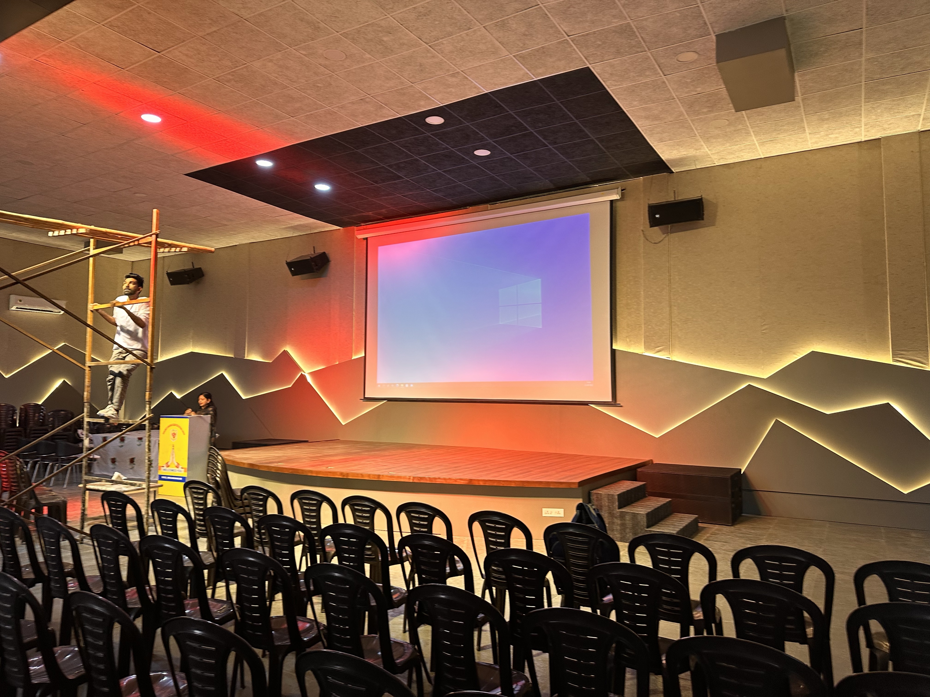 School hall with a projection screen over the stage, fabric-panelled walls with lit recesses and wall-mounted loudspeakers